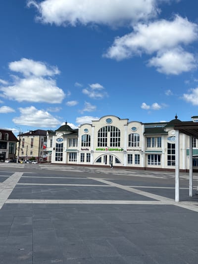 The image depicts a sunny day in Noginsk, Russia, featuring a modern building with businesses on the ground floor, including a pharmacy and a clothing store. The building has a cream-colored facade with large windows and a green roof. In front of the building is a spacious paved plaza with a geometric pattern of dark gray and lighter gray tiles. The sky is blue with scattered white clouds. Several cars are parked in the background, and a few pedestrians are visible on the plaza and near the building. The overall scene is one of urban activity and architecture on a clear day.
