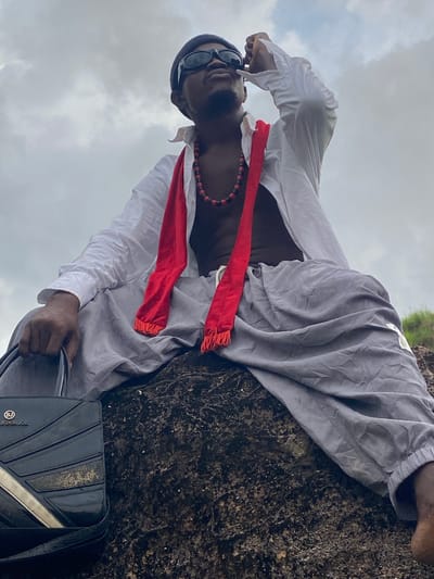 The image, taken in Nguzu Edda, Nigeria, features a dark-skinned male positioned outdoors against a cloudy sky. He is leaning or lying horizontally on a dark, rough, textured surface, which appears to be a rock formation.
The man is dressed in a white long-sleeved shirt, unbuttoned at the front, revealing a red and black beaded necklace. A bright red scarf or sash, with fringed ends, is draped around his neck and chest. He wears loose-fitting grey trousers. His head is covered by a dark beanie or cap, and he wears black sunglasses.
His right hand holds a black shoulder bag with a distinct design, featuring geometric ridges and a metallic diagonal stripe. Visible text on the bag includes "LONDON JOE" and a circular "BJ" logo. The background is composed solely of a grey, overcast sky, indicating a cloudy daytime setting. The individual appears to be in a relaxed pose, possibly resting or posing for the camera in a natural environment.