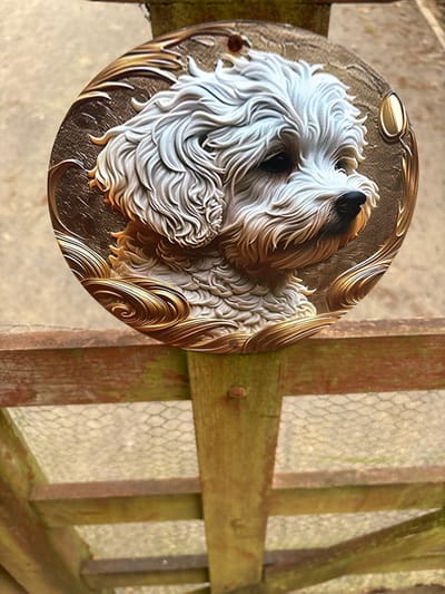 A circular, relief-sculpted plaque depicting a white, fluffy dog's head and upper body is centered vertically. The dog, likely a Maltese or similar breed, faces slightly right, rendered with intricate detail in its fur. The plaque's background features textured bronze tones with gold-colored swirling decorative patterns. A small hole is visible at the top edge of the plaque. This plaque rests against a weathered wooden structure, appearing to be part of a fence or gate. The wooden structure consists of horizontal reddish-brown beams and a vertical post, all showing signs of weathering and some green discoloration. A nail head is visible in the vertical post. The blurred background above the wooden structure is a light-colored ground surface. Below the main horizontal wooden beam, a section of hexagonal wire mesh is visible, consistent with chicken wire, spanning horizontally above a blurred ground surface. The scene is located in Snailbeach, United Kingdom.