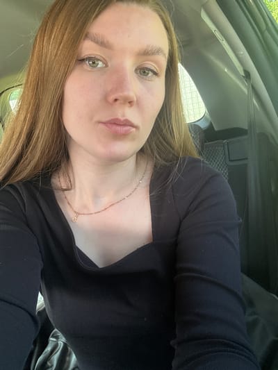 A young woman is in the interior of a vehicle, possibly a car, as evidenced by the dashboard, window, and seat visible. She is wearing a black, long-sleeved garment. She has light brown hair and fair skin. She is wearing a delicate necklace with a pendant. The location is not identifiable as Италмас, Russia from this image.