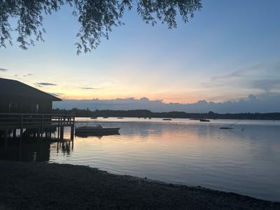 The image depicts a serene lakeside scene at dusk in Rimsting, Germany. The sky is a gradient of soft blues, transitioning to hues of orange and pink near the horizon, suggesting a sunset or sunrise. Wispy clouds are scattered across the sky, and the silhouette of trees and buildings forms a dark line along the distant shoreline.
In the foreground, a dark, pebbly shore meets the calm water. To the left, a wooden structure, possibly a boathouse or pier, extends over the water, supported by pilings. Several people are visible on the pier, casting shadows on the water. A small white motorboat is docked beside the pier.
The lake is dotted with several sailboats and other vessels, some anchored and others appearing to be in motion. The water reflects the colors of the sky and the structures, creating a peaceful, mirrored effect. The overall atmosphere is tranquil and natural, capturing the beauty of a lakeside evening in Rimsting.