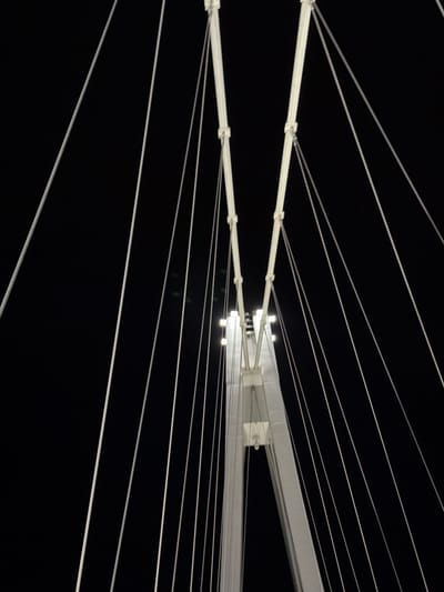The image presents a stark, abstract composition dominated by white lines against a completely black background. The primary subjects are a series of white, taut lines or wires, some appearing as thin strands and others as thicker, horizontal bars. These lines are arranged in a geometric pattern, suggesting a clothesline or an outdoor drying rack structure.
In the middle-left, two prominent white horizontal bars are visible, connected by small clips from which several thinner white lines extend. These lines radiate outwards, both upwards and downwards, and further to the right, creating a sense of depth and perspective. To the right side of the image, a more complex, lighter-colored structural element, possibly metallic or plastic, is visible. This structure incorporates several small, bright white lights that cast subtle illumination on the surrounding lines and surfaces.
The entirely black background indicates that the photograph was taken at night or in a very dark environment, with the white objects illuminated by artificial light sources embedded within the structure. There are no people visible, and no specific activity or event is taking place. The scene is devoid of any discernable weather conditions, and no textual elements are present. While the location context provided is Osijek, Croatia, there are no visual cues within the image itself to confirm this. The overall impression is one of minimalist geometry and strong contrast.