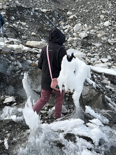 The image presents an overhead view of a person traversing a rugged, icy landscape, characteristic of a high-altitude glacial or periglacial environment, likely near Khumjung, Nepal. The subject is wearing a black hooded jacket, maroon-colored trousers, and a light pink crossbody bag slung over their shoulder. Their hood is up, covering their head.
The ground is a mixture of dark grey and black gravel, punctuated by numerous light grey and white rocks of varying sizes. Patches of white ice and fragmented sheets of ice are scattered across the terrain, creating an uneven and potentially slippery surface. A large, irregularly shaped piece of ice with a distinctive hole in its center is positioned near the person's left side, while other ice formations are visible to the right and in the foreground.
The person appears to be carefully walking or standing on this mixed rock and ice ground. A thin blue rope is visible in the lower left corner, extending out of the frame, which might indicate they are roped up for safety, common in glacier travel or challenging treks. The scene is illuminated by bright, even light, suggesting daytime. The small text on the pink crossbody bag is visible but too indistinct to be clearly read. The overall impression is one of cold-weather trekking in a stark, mountainous setting.