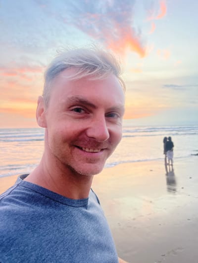 The image depicts a beach scene at either sunrise or sunset in Kerobokan Kelod, Indonesia. In the foreground, a light-skinned male with short, light-blonde hair and light stubble is centered on the left, smiling directly towards the camera. He is wearing a blue crew-neck t-shirt. The background features a wide expanse of ocean with gentle waves breaking on a sandy shore. The sky above is characterized by light blue, orange, and pink hues from scattered clouds, indicating early morning or late evening light. On the distant wet sand, two heavily blurred and silhouetted figures are standing close together, their reflections visible on the reflective surface of the sand. No other specific objects or distinct interactions are discernible beyond these elements.