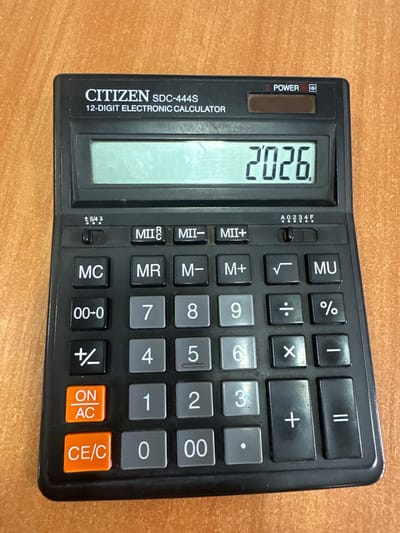 This is a top-down, eye-level, medium shot of a black Citizen SDC-444S 12-digit electronic calculator resting on a wooden surface. The calculator is oriented vertically, with the screen at the top. The screen displays the number "2026" in a large, clear font. To the left of the screen, there is a "POWER" button and a small slider switch labeled "5/4". The calculator has a standard layout of buttons, including number keys, mathematical operation symbols (+, -, x, ÷), memory functions (M+, M-, MR, MC), percentage (%), square root (√), and an "ON/AC" and "CE/C" button. The setting appears to be indoors, given the presence of the calculator and the wooden desk. There are no people or animals visible. The lighting suggests it could be daytime, but the exact time of day and weather are not discernible. The overall impression is a static product shot of a common office or personal electronic device.