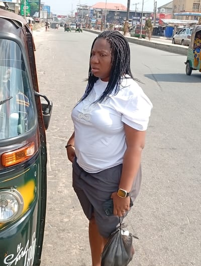 The image captures a daytime street scene in Aba, Abia, Nigeria. An adult female, with dark skin and long, dark braided hair, stands upright on an asphalt road surface. She wears a white short-sleeved t-shirt featuring a light-colored design with text, and dark grey shorts. A gold-colored watch with a dark strap is present on her left wrist, and a ring is on her left ring finger. She holds a black smartphone and a black plastic bag in her left hand; her right hand is placed into her shorts pocket. Her facial expression shows a slight openness of the mouth, and her gaze is directed towards her right (the viewer's left).
To her immediate left, the partial front-left section of a green and yellow motorized tricycle (keke Napep) is visible, showing a headlight, an amber indicator light, and part of the windshield.
The background features a wide asphalt road extending into the distance, populated by multiple tricycles and other vehicles. Commercial buildings line both sides of the street, displaying various signboards, including visible text such as "CHURCH HILL BANK" and "FIRST BANK." Utility poles with overhead wires are visible along the right side of the road, which also has a concrete curb or median. A large billboard with green branding is situated on the far left in the distance. The sky above is clear.