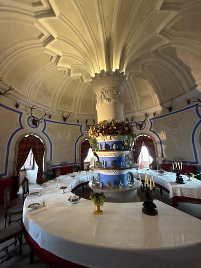 The image captures a room in the Palácio Nacional de Queluz in Sintra, Portugal. The room features a long, curved dining table set with plates, silverware, and decorative items like candelabras and floral arrangements. A central, multi-tiered decorative centerpiece with flowers and plates dominates the table. The architecture includes a dome-shaped ceiling with radial detailing emanating from the center. Mounted antlers decorate the walls, along with decorative carvings. Curtained windows are visible around the room. There are no people visible in the frame.