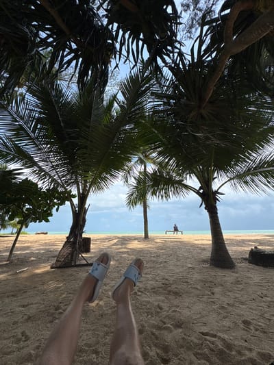The image depicts a tropical beach scene in Ban Khuk Khak, Thailand. The foreground shows a person's legs and feet, wearing light-colored slides, resting on the sand. The perspective is from the ground looking up and out towards the ocean. Several palm trees with broad fronds dominate the midground, casting dappled shadows on the sandy beach. In the distance, a lone individual sits on a wooden bench facing the turquoise ocean and a slightly overcast sky. The environment suggests a relaxed, seaside atmosphere.