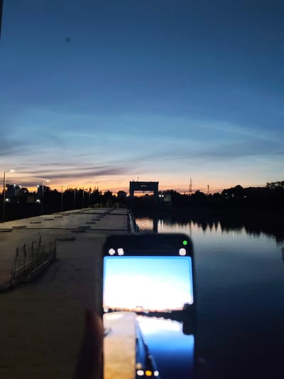 The media file is a photograph taken at dusk. The foreground shows a person holding a smartphone, capturing the scene. The smartphone screen displays a bright, almost blown-out image of a sunrise or sunset, reflecting in the water. In the background, a bridge or walkway extends across a body of water. Streetlights are visible along the walkway, and trees and buildings are silhouetted against the colorful twilight sky. A power line tower can also be seen in the distance. The overall mood is peaceful and serene, with the warm hues of the sunset contrasting with the deepening blues of the sky and water. The setting appears to be an urban or semi-urban environment near a body of water, possibly a river or canal. There is no visible text in the image.