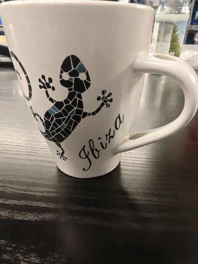 A white ceramic mug with a black and blue mosaic gecko design and the word "Ibiza" in black script is placed on a dark, textured wooden surface. The mug is positioned at an angle, showing its handle and a portion of its interior. In the background, out of focus, are items on a shelf including a clear bottle with liquid and greenery, possibly a plant, and some white packaging. The lighting suggests an indoor environment. The location of Hofgeismar, Germany is not discernible from the image.