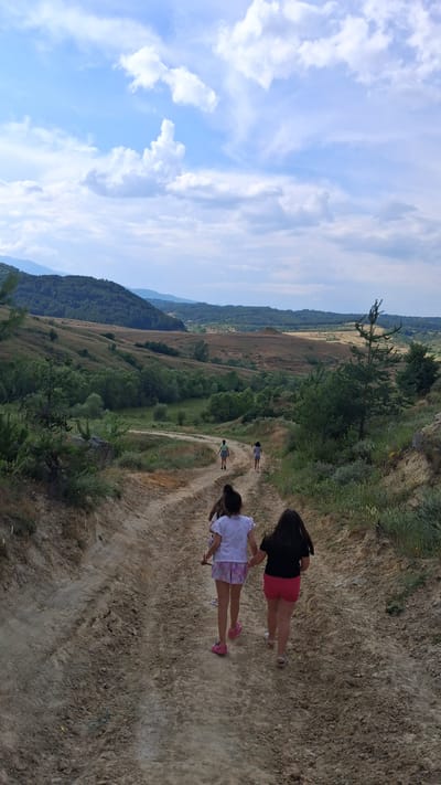 The image depicts a scenic, rural landscape in Dobrotino, Bulgaria. The foreground features a dusty dirt path winding along a hillside, with sparse vegetation and dry, light-colored soil. On the path, two young girls are walking away from the camera. The girl closer to the viewer is wearing a white shirt with a colorful patterned skirt and pink sneakers. The girl behind her is wearing a black t-shirt and pink shorts. Further down the path, two more figures are visible, seemingly walking in the same direction.

The middle ground reveals a lush, green valley with rolling hills covered in trees and vegetation. The landscape recedes into the distance, with layers of hills under a bright, cloudy blue sky. A single tree stands prominently on the edge of the path in the mid-ground. The overall impression is one of a sunny day, likely late morning or early afternoon, given the bright light and clear sky. The scene evokes a sense of leisure and outdoor activity, possibly a hike or a stroll through the countryside. There is no visible text in the image.