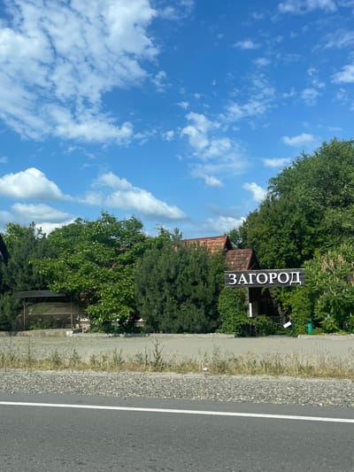 A vertical image shows a street scene in Donskoe, Russia, on a sunny day with a bright blue sky and scattered white clouds. On the right side of the image is a paved road with a white line marking the edge. Adjacent to the road is a gravel shoulder, followed by a paved pedestrian walkway or sidewalk. Beyond the walkway, there is lush green foliage and trees, interspersed with a few buildings with terracotta roofs.
A notable object in the scene is a dark wooden sign with white Cyrillic text that reads "ЗАГОРОД," meaning "out of town" or "suburb." The sign is placed amongst the trees, indicating the edge of a built-up area or perhaps an entrance to a residential community. The overall atmosphere is peaceful and suggests a rural or semi-rural setting. The lighting indicates it is daytime, likely mid-morning to late afternoon. There are no people visible in the image, and no specific activity is taking place. The focus is on the natural elements of trees and sky, combined with the man-made elements of the road and the directional sign.