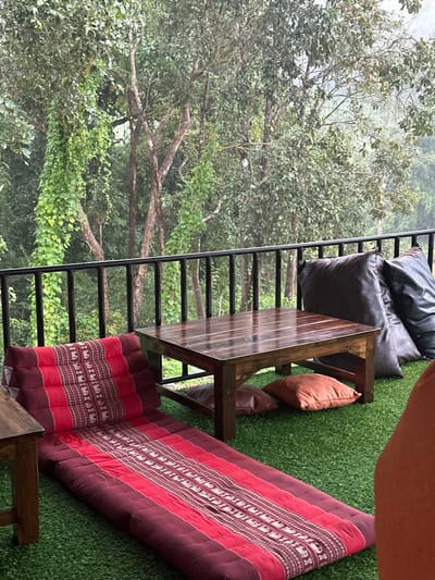 The image depicts an outdoor seating area, likely a balcony or elevated terrace, situated in Ban Paem Bok, Thailand, overlooking a dense, verdant forest. The ground surface of the seating area is covered with artificial green turf. A black metal railing separates the foreground from the forested background, which consists of numerous trees, some covered in vines, under what appears to be an overcast or misty sky.
Within the seating area, a rectangular wooden coffee table with a dark, polished surface occupies the center-right. Below the table, two square floor cushions, one brown and one orange, are positioned on the turf. To the right of the table, two large, dark-colored beanbag cushions, one dark gray and one black, are stacked against the railing. To the left of the main table, a long, traditional Thai-style folding mat with an elevated triangular headrest is laid out. This mat features a distinct pattern of red and maroon stripes interspersed with white elephant motifs. A portion of another wooden table is visible on the far left. A human arm, wearing an orange-brown garment, is partially visible on the far right edge of the frame, indicating the presence of an individual in the scene. No explicit actions are observed beyond the arrangement of the lounge area.