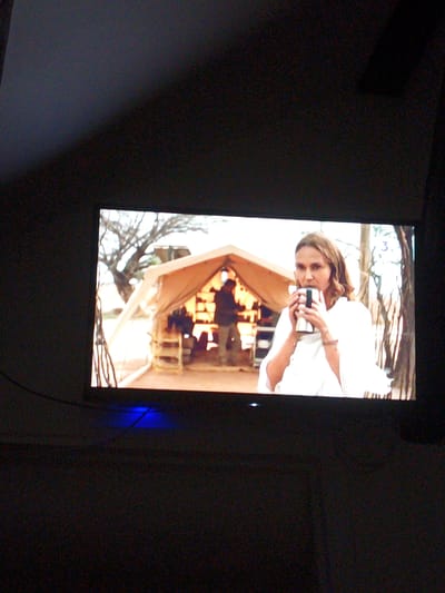 The image shows a woman with medium-length brown hair, wearing a white garment, holding a silver mug to her lips. She is standing in front of a tent. Behind her, a person in a dark shirt and pants is visible inside the tent. The tent is beige and has an open front. There are wooden steps leading up to the tent. In the background, there are trees with bare branches and a hint of blue sky. A small blue light is visible at the bottom of the image. There is no discernible information within the image that suggests it is located in Montgeron, France.