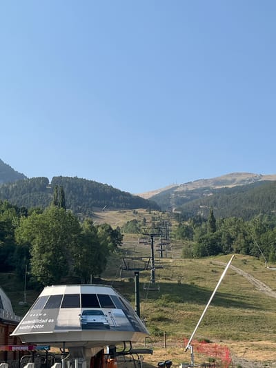 This is an outdoor, eye-level shot of a ski lift in the Pyrenees mountains near Benasque, Spain. The sky is clear and blue. In the foreground, a portion of a ski lift station is visible, adorned with a large advertisement featuring a silver car and text that reads "VOLVO sostenibilidad es nuestro motor." Adjacent to the station, ski lift chairs are suspended on a cable, extending up a grassy mountainside. The mountain slopes are covered in a mix of trees and sparse vegetation, with visible ski trails and more ski lift towers ascending towards the distant peaks. The overall scene depicts a mountain resort during the off-season, with the ski infrastructure prominently featured against a backdrop of natural landscape.