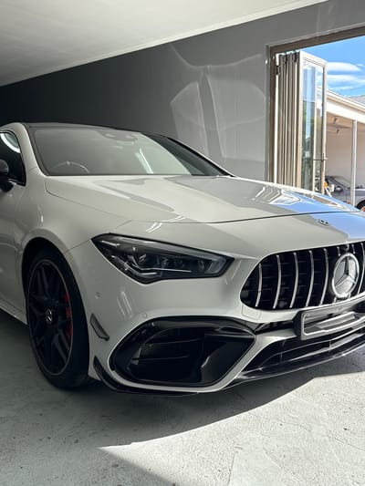 A low-angle, close-up photograph captures the front-three-quarter view of a pristine white Mercedes-Benz AMG car, likely parked indoors or under a covered area in Blouberg, South Africa. The car's glossy white bodywork reflects the bright blue sky with subtle cloud formations, along with a modern building featuring glass panels and balconies, suggesting an open or windowed overhead structure.
The dominant feature is the car's aggressive front fascia, highlighted by the prominent Panamericana-style grille with vertical chrome slats and the iconic Mercedes-Benz three-pointed star logo at its center. Sleek, dark LED headlights are visible, flanking the grille, and the front bumper incorporates black aerodynamic elements and grilles. Below, black multi-spoke alloy wheels are visible, revealing vibrant red brake calipers, indicating high-performance specifications.
The setting includes a textured, light-colored wall or ground surface on the right side of the vehicle, where a distinct shadow is cast by the car, suggesting strong overhead sunlight. On the left, a plain, light-colored wall runs alongside the vehicle. The car is stationary, and no people are visible in the frame. The image conveys a sense of luxury and powerful automotive design under clear, sunny weather conditions.