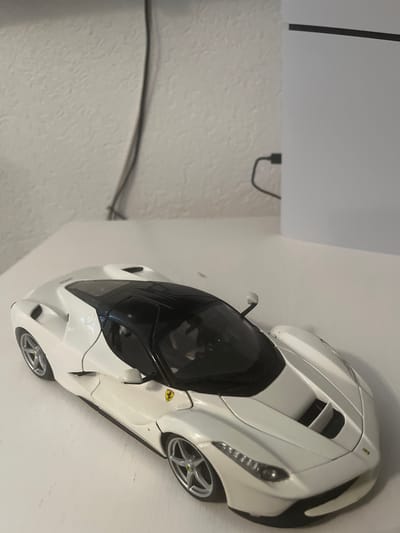 A white and black toy Ferrari is displayed on a white shelf. The car is a model of a modern sports car, with a sleek design and prominent vents on the hood. The interior appears to be black, and the wheels are silver with five spokes. A small yellow Ferrari logo is visible on the side of the car. The background consists of a textured white wall and a portion of a white shelf or table. A black cable is visible on the textured wall, extending from the top left corner of the image and curving downwards. The lighting suggests an indoor setting, possibly during the day with artificial light. No people are visible in the image. The overall scene depicts a static display of a collectible toy car.