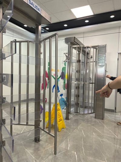 A slightly tilted shot captures a stainless steel, security barrier-style gate in a modern interior with gray marble flooring. The gate features a revolving metal bar section and clear acrylic panels. A hand points toward the barrier. "Salida" is visible on a sign above the gate. The background includes colorful children's decorations and yellow "Caution Wet Floor" signs placed haphazardly near the gate. The overall impression is of a public space exit.