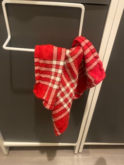 The image displays an indoor scene featuring a red and white plaid textile, identified as a kitchen towel, folded and draped over a white, U-shaped metal towel rack. The towel exhibits a pattern of broad red bands intersected by narrower white lines, forming a grid, with some areas featuring finer red and white checks. The towel rack is mounted on or positioned against a dark gray, smooth vertical surface, possibly a cabinet door or appliance panel. To the right of the towel and rack, a white vertical structural element, likely a frame or support, extends from the lower portion of the image upwards. The floor beneath is a light gray or beige tone. No individuals are present in the image. The visual content does not provide identifiable details regarding a specific location within the city of Oberhausbergen, France.