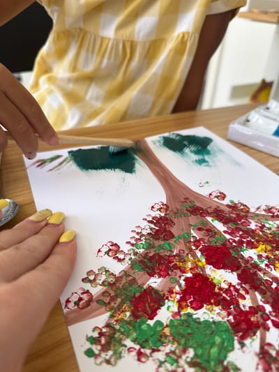 In an indoor setting, two individuals are engaged in painting on a white sheet of paper placed on a wooden table. The person in the foreground, with yellow-painted fingernails, is working on the lower portion of the painting, which depicts a tree with red and green splotches representing leaves and flowers. They are also seen holding a spoon. To the left, a hand is holding a wooden-handled brush, applying green paint to the upper part of the tree, presumably for its foliage. In the background, a person wearing a yellow and white checkered dress is visible. A paint tray containing blue and white paint is situated to the right of the painting. The environment suggests an art activity. The location of Khan Chbar Ampov, Cambodia is not discernible from the image.