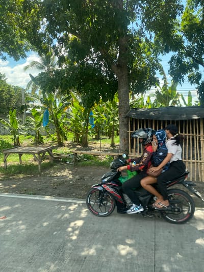 Three individuals are riding on a motorcycle on a paved road in a tropical environment, likely Koronadal City, Philippines, characterized by lush vegetation including banana trees. The rider in front is wearing a helmet and a colorful jacket. Seated behind the rider are two individuals, one with a blue headscarf, and a third person at the very back. They appear to be traveling alongside a roadside area with a makeshift bamboo fence and some wooden tables. The scene is illuminated by natural daylight, with shadows indicating a sunny day.