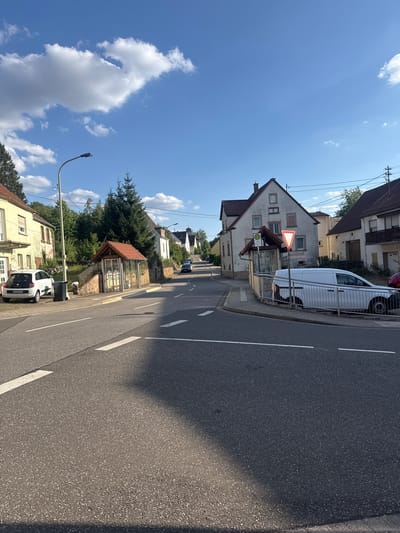 The image depicts a street scene in Hüffler, Germany, under a bright blue sky with scattered white clouds. The street is paved and has white lane markings. On the left side of the road, a yellow house with a white trim and shutters is visible, with a white Renault Twingo parked in front of it next to a black trash bin. Further down the sidewalk is a small bus stop with a red-tiled roof and glass panels. Across the street, a white van is parked behind a metal fence. Behind the van and fence, a white building with a red-tiled roof and multiple windows is prominent. Several other houses and trees line the street, which curves gently into the distance. A traffic sign with a red border and white triangle is also visible near the bus stop. The lighting suggests it is daytime, likely late afternoon due to the shadows.