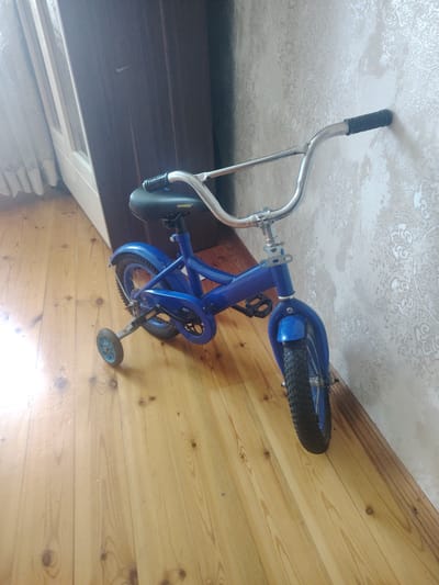 A blue children's bicycle with training wheels stands upright, leaning against a textured wall. The bicycle has a chrome handlebar with black grips, a black seat, and blue fenders. The main frame of the bicycle is also blue. The training wheels are visible on the rear of the bike, one on each side. The bicycle is positioned on a light-colored wooden floor. In the background, to the left, a portion of a door or cabinet with white trim and a reflective surface is visible, with sheer curtains covering a window. The wall behind the bicycle has a repeating pattern in a lighter color. The lighting suggests it is daytime. The scene appears to be indoors, likely a room in a residential setting. There is no discernible activity taking place; the bicycle is stationary. Some text is visible on the seat, appearing to read "mongoose." The location context is Hovsan, Azerbaijan.