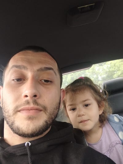 A man and a young girl are in a car. The man, likely a father, is taking a selfie with the girl. The man has a beard, dark hair, and is wearing a black hooded sweatshirt. The girl has curly brown hair, a pink hair clip, and is wearing a light purple shirt with a polka-dot jacket. The car interior is visible, as is greenery visible through the car window. No specific information indicates the exact location within Stafford, United Kingdom.