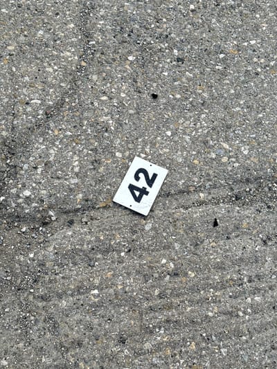 A white rectangular marker with the number "42" in black, bold font lies on a textured, gray asphalt surface. The asphalt is speckled with various sizes of gray and white pebbles, with some darker aggregate visible. The marker is positioned at an angle, with the top left corner angled slightly upwards and to the left, and the bottom right corner angled downwards and to the right. Shadows are cast faintly behind and to the right of the marker, indicating a light source from the upper left. The surface shows subtle signs of wear, with some faint darker lines and indentations suggesting a well-trafficked area. This scene was captured in Fontevivo, Italy.