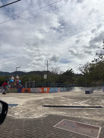 The image shows an empty skate park in San Rafael, Costa Rica. The skate park is located at 9.938 latitude and -84.153 longitude, surrounded by trees and hills. The park is made of concrete and has a ramp. There is a playground in the background. There are no people in the image. The sky is cloudy.  The skate park is surrounded by a fence and has a  metal grate drain in the middle. The park is closed. The ramp is empty.  The playground is made of metal and plastic and has a colorful paint job. It is surrounded by a fence and a net. There are no people on the playground either. There are wires running across the sky. The area is surrounded by green vegetation. There are no cars in the picture.  A  piece of a rearview mirror is in the lower left corner of the image. The ground is covered in a brick pattern.  The image was taken during the day.