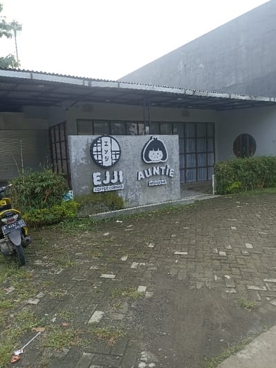 The image shows the exterior of a commercial building located at coordinates -7.418, 109.251 in Purwokerto, Indonesia. The building appears to be a cafe or restaurant with two businesses: "Ejji Coffee Corner" and "Auntie Noodle Bar." The building is constructed of grey concrete blocks and has a black awning above the entrance. The businesses are marked with signs featuring stylized graphics and the names written in black lettering on a white background. The businesses are located on the side of the building and face a paved area composed of grey brick-like pavers. A small, yellow motorcycle is parked on the pavers in front of the building. Green bushes surround the building, and a portion of a white plastic bag is visible on the ground in the lower left corner of the image. The sky is overcast and grey.