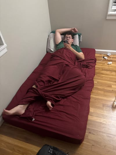 A young man with short dark hair and glasses is depicted lying on his left side on a maroon-colored futon or mattress. He is wearing a green t-shirt and is absorbed in looking at a smartphone, which he holds in his right hand. His head rests on a white pillow, and most of his body is covered by a matching maroon blanket or sheet. One bare foot is visible, peeking out from under the blanket towards the lower right of the frame.
The setting is an indoor room with light grey walls and light-toned hardwood flooring. A white window frame is partially visible in the upper left corner of the image. Above the floor and near the head of the futon, multiple white electrical outlets and tangled white wires are visible on the wall. A section of a dark grey or black bag can be seen on the floor to the right. The scene conveys a sense of casual relaxation, likely in a residential space in Franklin Township, USA. The time of day is not overtly clear, but the indoor lighting suggests a comfortable, unhurried atmosphere. No discernible text is visible in the image.