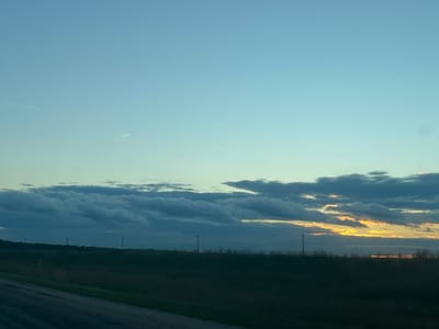 The image depicts a rural landscape at dusk, featuring a vast expanse of sky transitioning from a pale blue to a soft yellow near the horizon. A thin, wispy contrail from an airplane is visible in the upper left quadrant of the sky. The lower portion of the image is dominated by a dark, silhouetted horizon line, suggesting a field or grassland. Several utility poles or communication towers are sparsely scattered along this horizon. A dirt or gravel road occupies the bottom left corner of the frame, leading into the scene. The lighting suggests it is either sunrise or sunset, with the sun's rays reflecting off the lower edges of some of the scattered clouds, casting a warm, golden glow amidst the darker, cloud-laden sky. The overall impression is one of quiet solitude in an open, undeveloped area.