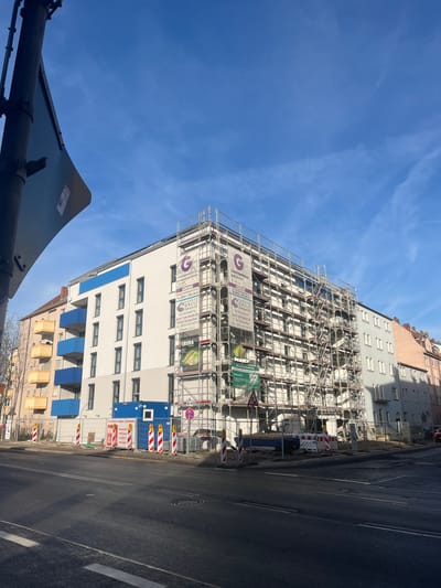The image depicts a vibrant street scene in Schweinfurt, Germany, centered on a multi-story building undergoing renovation, under a clear blue sky. The photograph is taken from a low, upward angle, causing the building and street to appear diagonally oriented.

The upper floors of the building are entirely enveloped in extensive metal scaffolding, signifying active construction or refurbishment. Attached to this scaffolding are several white banners featuring a prominent purple "G" logo and text that reads "GANTT GALLI GRUPPE." Further details on these banners include "Immobilien - Projekte - Neubau - Sanierung" (Real estate - Projects - New construction - Renovation), as well as an address "Ludwigstr. 24" and contact information such as "09721-64670-0" and "www.ganttgalli.de." Another green banner displays "ZEHA" alongside a graphic resembling a pear or apple.

Below the scaffolding, the lower section of the building presents a newly finished white facade with numerous dark-framed windows. Blue awnings are extended over some windows on the bottom floors. Other buildings are partially visible in the background and to the left of the main structure.

Adjacent to the building, a paved street runs parallel, featuring clear road markings and a manhole cover. Along the roadside, construction barriers with distinctive red and white stripes are erected, some bearing the German words "BAUSTELLE" (Construction Site) and "ACHTUNG BAUSTELLE" (Attention Construction Site). A standard "No Stopping" or "No Parking" traffic sign (red circle with a diagonal line) is also visible. In the lower-left foreground, a dark, angled traffic sign is partially captured.

The sky is a brilliant, cloudless blue with only a few faint, high-altitude streaks, indicating bright, sunny daylight. There are no people visible in the scene. The overall impression is one of an active urban development or renovation project.