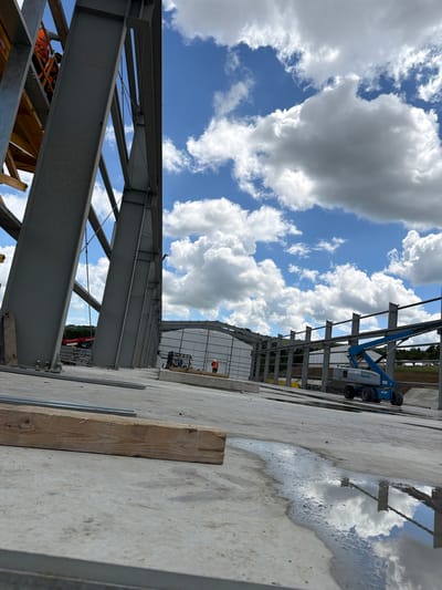 The image depicts a construction site in Skrzydlna, Poland, focusing on the skeletal structure of a building. Large steel beams, forming the framework of the structure, dominate the left side of the frame. In the background, a partially constructed building with a white facade and a row of vertical steel supports can be seen. A blue Genie S-85 boom lift is positioned near the structure, with its arm extended. A person wearing an orange safety vest is visible near the boom lift, and another individual in similar attire is standing further in the distance. The ground is concrete, with some areas showing puddles of water reflecting the cloudy sky. A wooden plank rests horizontally on the concrete in the foreground. The sky is blue with scattered white clouds.