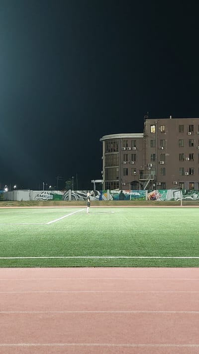 The image depicts an outdoor sports field at night, likely a soccer pitch, with a running track in the foreground. The field is illuminated, with white lines marking its boundaries. A lone figure, appearing to be a person in athletic attire, stands on the field. In the background, a multi-story building with numerous windows and air conditioning units is visible. The building has a distinctive rounded section. Behind the field and in front of the building, a mural or decorative wall runs along the perimeter, featuring various images and text, including the word "AMKUN." The sky above is dark. This scene is consistent with the sports complex located in Novocherkassk, Russia, a facility known for its athletic fields and surrounding infrastructure.