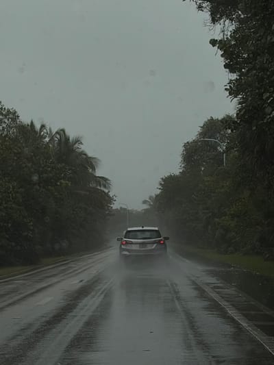 A white car drives on a wet road during heavy rain. Trees line the side of the road, and the sky is overcast and gray. Rain streaks across the image, obscuring details. The car's taillights are visible.