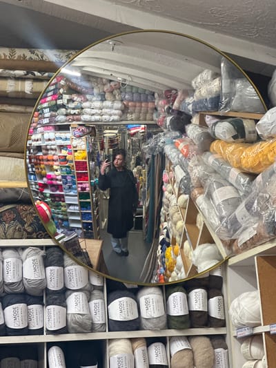 A woman is reflected in a gold-rimmed circular mirror surrounded by shelves of yarn. The mirror shows the interior of a yarn store. The shelves in the foreground contain rolls of yarn with labels. A woman in a black coat is taking a selfie in the reflection. The shelves reflected in the mirror display a wide range of yarn colors. Some fabric rolls are visible below the shelves in the mirror's reflection. The yarn labels indicate the store is in Sevenoaks, United Kingdom, selling "MODE at ROWAN" yarn.