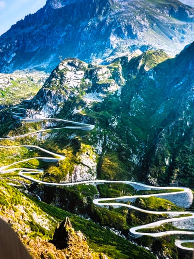 This image captures the dramatic switchbacks of the Susten Pass road in the Swiss Alps, near Schattdorf, Switzerland. The winding road, constructed with stone retaining walls, carves its way up the steep, verdant mountainside, illuminated by the sun. Patches of green grass and vegetation cover the rocky slopes, interspersed with stark white rock formations. In the distance, imposing, deep blue mountains rise, their peaks shrouded in shadow. The sky above is a bright, clear blue. There are no people or vehicles visible in the scene, emphasizing the grandeur and isolation of the natural landscape and the engineering feat of the road. A small, dark-colored structure, possibly a building, is visible nestled amongst the greenery along the road in the mid-ground.