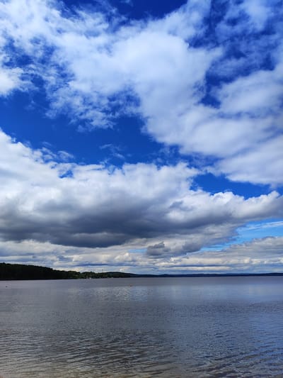 A wide shot captures a serene lake with a forested shoreline under a dynamic sky filled with both bright blue patches and dark, dramatic clouds. The water's surface is gently rippled, reflecting the muted colors of the sky. The shoreline is densely wooded with dark green trees, and in the distance, there are hints of structures and a faint silhouette of the land. Two small orange buoys are visible in the water. The overall impression is one of a tranquil, natural landscape on a day with changing weather conditions. This scene is consistent with locations around the city of Snezhinsk, Russia, known for its numerous lakes and forested areas.