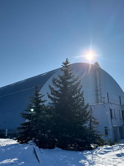 The image displays a winter scene at the Chernobyl Nuclear Power Plant, located within the Chernobyl Exclusion Zone, Ukraine. While not situated directly within the city limits of Pryp'yat', the plant is approximately 15 kilometers northwest of Pryp'yat', with both locations being integral parts of the Exclusion Zone.

The central subject is the New Safe Confinement (NSC), a massive, arched, blue-grey metallic structure covering Reactor Unit 4. The NSC occupies the midground and background, exhibiting some snow accumulation on its upper surface. A prominent vertical utility structure, appearing as a ladder or maintenance access, is affixed to the right side of the NSC.

In the foreground, several dark green evergreen trees, dusted with snow, partially obscure the base of the NSC and an adjacent, smaller, light-colored building. This ancillary building, situated to the right of the NSC, features multiple rectangular windows and what appears to be an entrance. Barbed wire is visible along the top of a fence or low wall associated with this building. The ground is uniformly covered in white snow, displaying some visible darker patches and faint tracks. A partial view of a circular white traffic sign with a red border and a black horizontal line is visible on the far left. The sky is clear and bright blue, with the sun positioned in the upper right quadrant, emitting a strong lens flare. No individuals or active movements are visible.