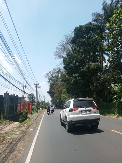 The image shows a street scene in daylight. A white SUV is in the foreground, positioned on the right side of the road. Two motorcycles are visible in the distance, also traveling on the right side of the road. The road has a solid yellow line marking the edge.

On the left side of the road, there is a fence and utility poles with numerous wires running along them. On the right side of the road, there are trees and other vegetation. The sky is blue with some clouds. There is no evidence to confirm the location is Nawng Hkar, Myanmar.