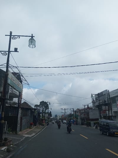 The image captures a daytime street scene in Thotagamuwa, Sri Lanka, under an overcast sky with patches of lighter clouds and some visible blue sky. A two-lane asphalt road, marked with a solid yellow center line and dashed yellow lane indicators, recedes into the distance. Numerous overhead electrical or telecommunication wires crisscross the upper portion of the frame.
On the left, a tall, dark, ornate street light pole with a cylindrical, ribbed lantern is prominent, accompanied by a smaller rectangular fixture. Behind this pole, a partially obscured, faded sign and a two-story building with a grey lower facade are visible. Further along the left, a single-story building with a brown roof and dark, weathered walls, some appearing graffiti-covered, borders a narrow concrete sidewalk.
Traffic on the road consists primarily of vehicles moving away from the viewer. At least four motorcycles, each with a helmeted rider, are visible in the middle of the road. On the right side, a dark blue minivan, possibly a Suzuki Carry, is also moving away from the viewer. Additional vehicles are discernible further down the street.
The right side of the street is lined with commercial buildings. A notable two-story structure features a grey facade with prominent white vertical elements and displays signs, including one with "TAMARAS" and another with "SELAMAT" partially legible. Similar ornamental street lights with decorative arches spanning the road are visible at intervals along both sides of the street. Distant trees are visible under the cloud cover.