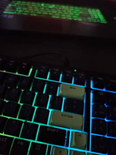 The image is a close-up, dimly lit, high-angle shot of a black mechanical keyboard with customizable RGB backlighting. The keys are black with white lettering and illuminated by streaks of green, teal, and blue light. Some keys are highlighted with a light green color, including the "ENTER" key and an adjacent key with an arrow pointing left. The top portion of the image shows a blurry reflection of more green-lit keyboard keys, possibly from a screen. The overall impression is that of a gaming or custom keyboard setup. There is no indication of the city of Versailles, France, or any specific location within it. No people or actions are depicted.