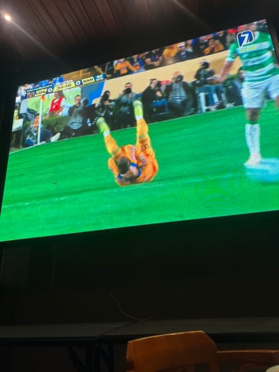 A television screen displays a soccer match. The score is tied 0-0 between UANL and BAN, with the game at the 34:46 mark. A player in orange lies prone on the green field, near the goal line. In the background, fans are seated and a player in green and white stands. The channel logo is visible in the top left corner. A wooden table is to the side.