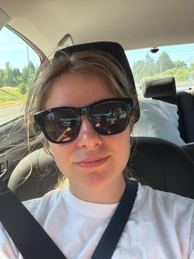 The image shows a young woman taking a selfie from the passenger seat of a car, looking towards the camera. She is wearing a white t-shirt and black sunglasses, with her blonde hair falling around her face. The car interior is visible, including the black headrest and a seatbelt across her chest. Through the car window, a bright, sunny daytime scene is visible, with green trees and a blue sky. The reflection in her sunglasses shows a partial view of the car's interior and the outside scenery. The image appears to have been taken outdoors on a clear day, likely in a suburban or rural area given the greenery. The woman's expression is relaxed and casual. There is no visible text in the image. The location context is Vancouver, United States.