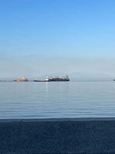 The image depicts a wide expanse of calm, blue water, reflecting the clear sky. In the distance, several large cargo ships are visible, traversing the horizon. The closest ship, on the right, has "MSC" clearly visible on its hull. Another cargo ship, painted with yellow and red containers, is further to the left. A smaller, lighter-colored vessel is positioned between these two larger ships. The background features a hazy, mountainous landscape, suggesting a coastal or port setting. In the foreground, a dark, textured surface, likely a promenade or walkway, runs horizontally across the bottom of the frame. The overall impression is one of a serene, maritime scene in or near the city of Thessaloniki, Greece, likely taken from its waterfront.