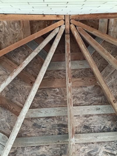 This is a close-up, low-angle shot looking up at the underside of a wooden roof structure, likely under construction or renovation. The main subjects are the wooden beams and OSB (Oriented Strand Board) sheathing that form the roof's framework. Several diagonal wooden rafters radiate from a central point, with some appearing weathered and painted white, while others are raw wood. Horizontal wooden planks are also visible, supporting the OSB. The OSB itself has a distinctive textured pattern.

At the very top of the image, a strip of white brickwork is visible, suggesting this structure is attached to a building. The lighting is diffused, indicating it's daytime, and there are no specific weather cues. The scene appears to be an outdoor construction site or the unfinished interior of a structure like a shed, gazebo, or the eaves of a house.

There is visible text on the OSB panels. Some legible parts include "ALLATION," "RDANCE WITH," "OVE ENDATIONS," "UCT SUPPORT 620-7400," "GE PANELS," "CHAT ENDS," "CHAT EDGES," "SIDE DOWN," "LADO ABA," "EN DESSOUS," "IMRECTION," "ENOTAXIS," "TONER SUPPORT -648-6893," "CANADA," and "OSB." These appear to be manufacturer markings or installation instructions. No people are visible in the image. The overall impression is one of raw construction materials and structural elements.