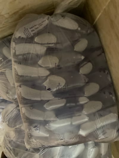 The image shows a close-up, angled view of several transparent plastic bags filled with individually wrapped, elongated, pale gray objects, possibly food items. These bags are stacked, with the foremost bag displaying text that includes "Batch No:", "Exp Date:", and "Mfg. Date:". The text is small and somewhat obscured by the plastic and the contents. The lighting is dim, and the background appears to be a textured, light brown surface, possibly wood or cardboard, suggesting the items are in storage or packaging. There are no people visible in the image. The location within the city of Dumnga, Nigeria, is not discernible from the visual information provided.