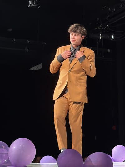 An indoor scene depicts a young male standing center on what appears to be a stage in Palos Verdes Estates, United States. The individual wears a two-button tan suit jacket over a dark grey collared shirt and a dark tie, with matching tan trousers. White socks are visible above his footwear, which is obscured by balloons. His hands are positioned near his chest, adjusting the lapels or collar of the suit jacket. He has short, dark brown hair and is looking slightly down and to his right, with a subtle smile on his face. The background is predominantly dark, indicating a theater or performance space, with overhead stage lighting fixtures and cabling visible. White numbers, including "79," "78," "77," "24," and "25," are affixed to structures in the dark background. The foreground features several inflated, translucent purple balloons scattered across the stage floor.
